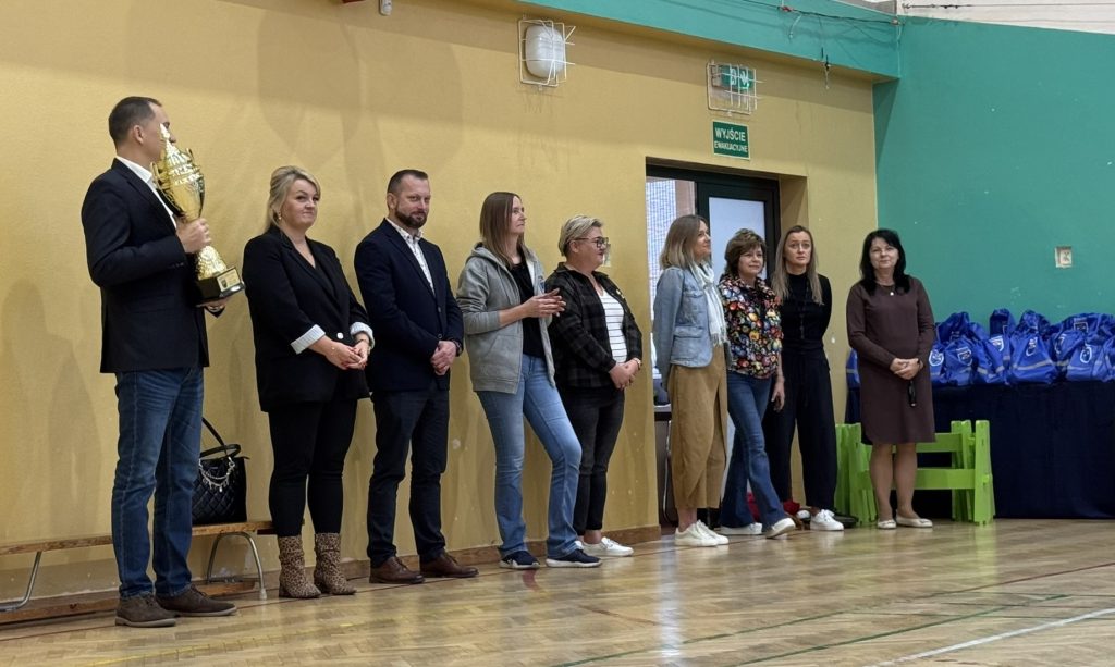 Przedstawiciele organizatorów i zaproszeni goście stoją w szeregu na sali gimnastycznej. Jeden z mężczyzn trzyma puchar, a za grupą widoczne są przygotowane niebieskie stroje sportowe.
