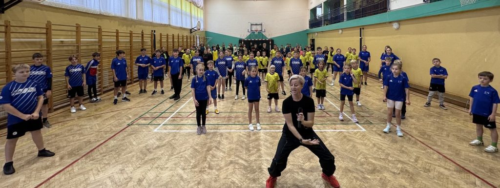 czestnicy wydarzenia sportowego stoją w kilku rzędach na sali gimnastycznej. Na pierwszym planie kobieta z mikrofonem zachęca do wspólnej aktywności.