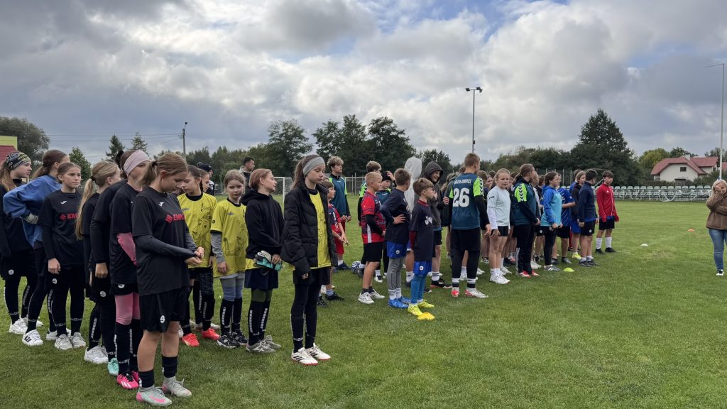 Grupa dziewcząt i chłopców w strojach sportowych ustawia się w szeregu na boisku trawiastym. W tle widoczne są bramki piłkarskie, ogrodzenie i drzewa.