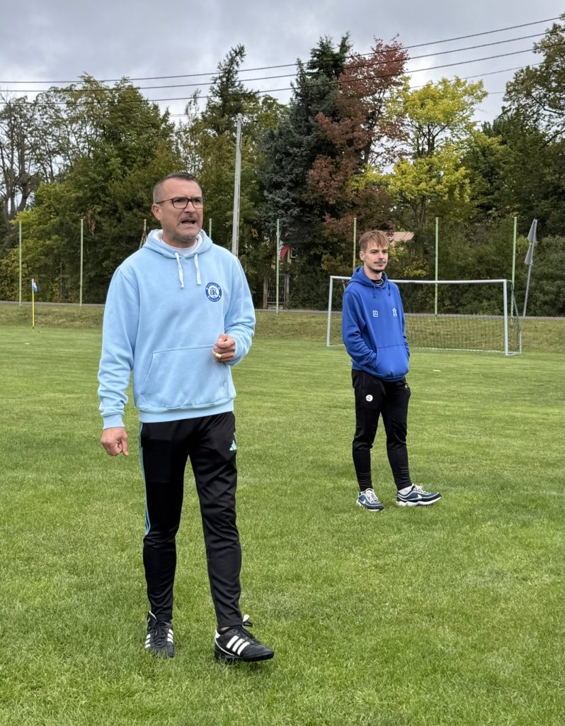 Dwóch mężczyzn w sportowych ubraniach stoi na boisku piłkarskim. Jeden z nich przemawia, a drugi obserwuje wydarzenie. W tle znajduje się bramka.