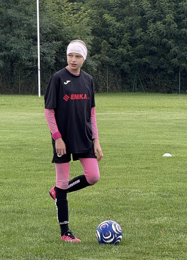 Młoda zawodniczka w czarno-różowym stroju sportowym i różowej opasce na głowie stoi na boisku obok piłki nożnej.