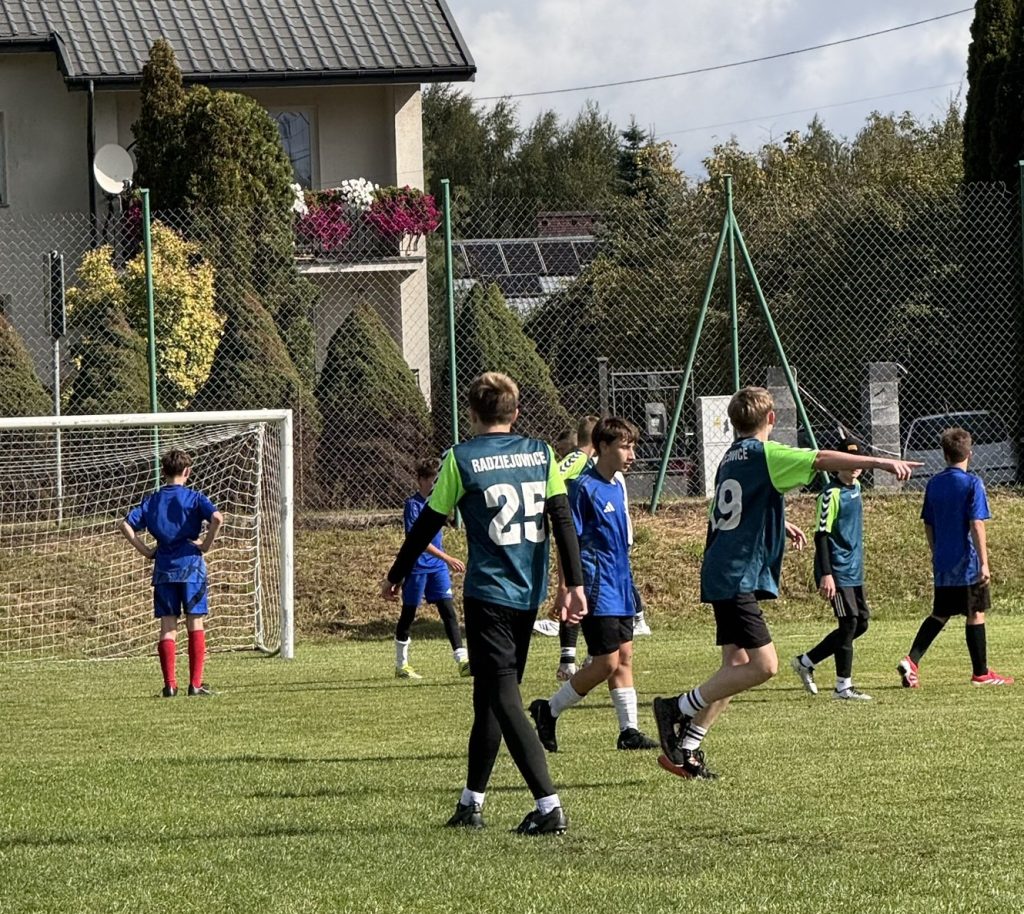Zawodnicy w niebieskich strojach sportowych rozgrywają ćwiczenie piłkarskie na boisku. Jeden z nich wskazuje kierunek podania, a pozostali poruszają się po murawie.