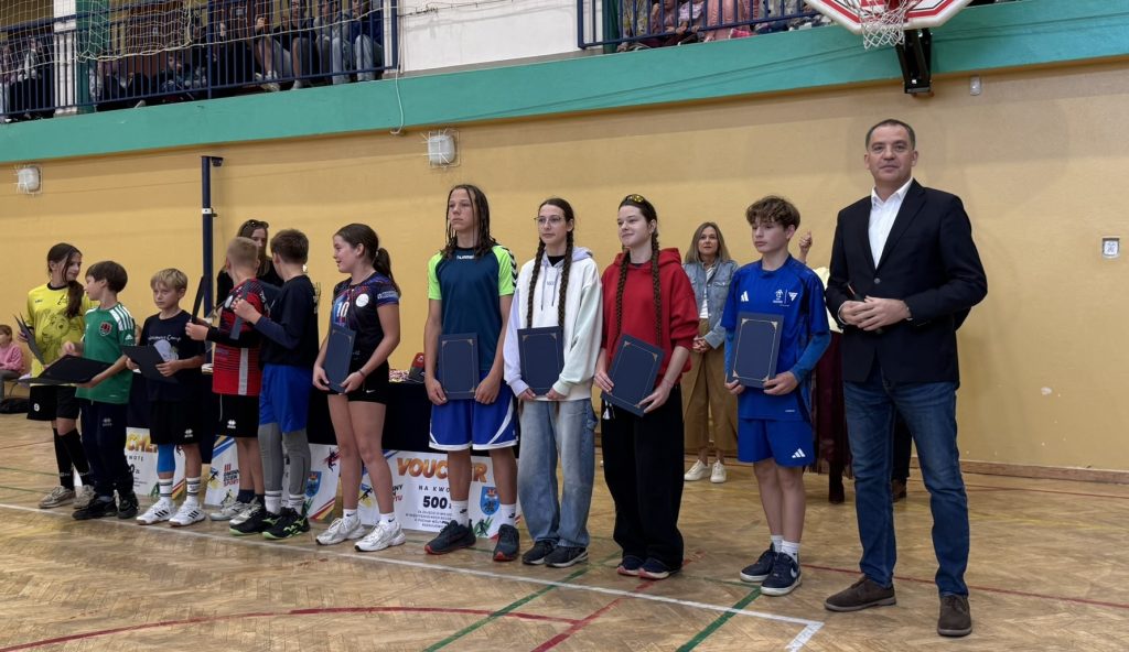 Grupa młodych sportowców prezentuje pamiątkowe dyplomy lub wyróżnienia podczas uroczystości na sali gimnastycznej. Obok uczestników stoi mężczyzna w garniturze.