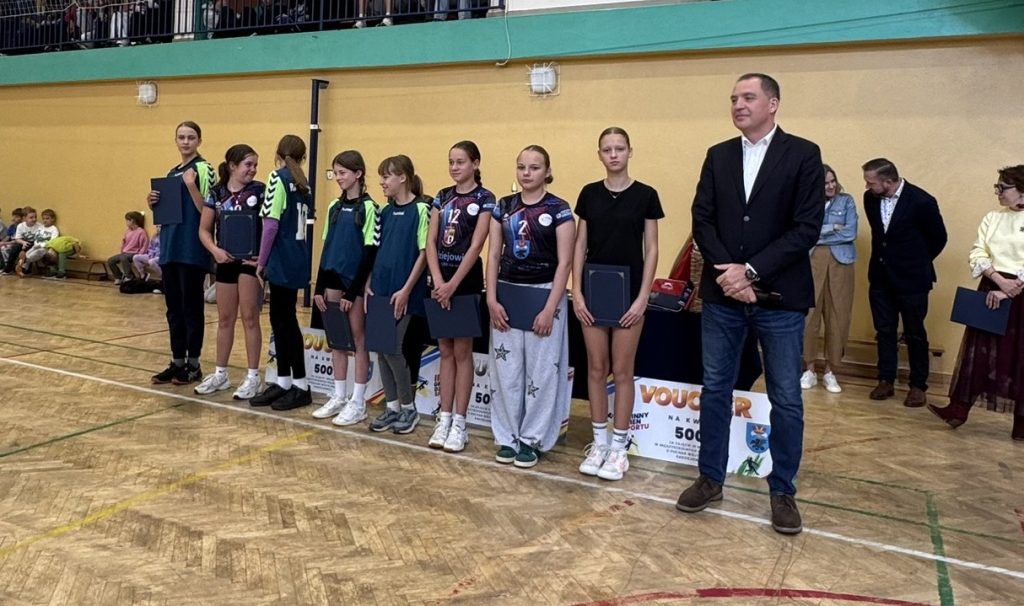 Grupa młodych sportowców prezentuje pamiątkowe dyplomy lub wyróżnienia podczas uroczystości na sali gimnastycznej. Obok uczestników stoi mężczyzna w garniturze.