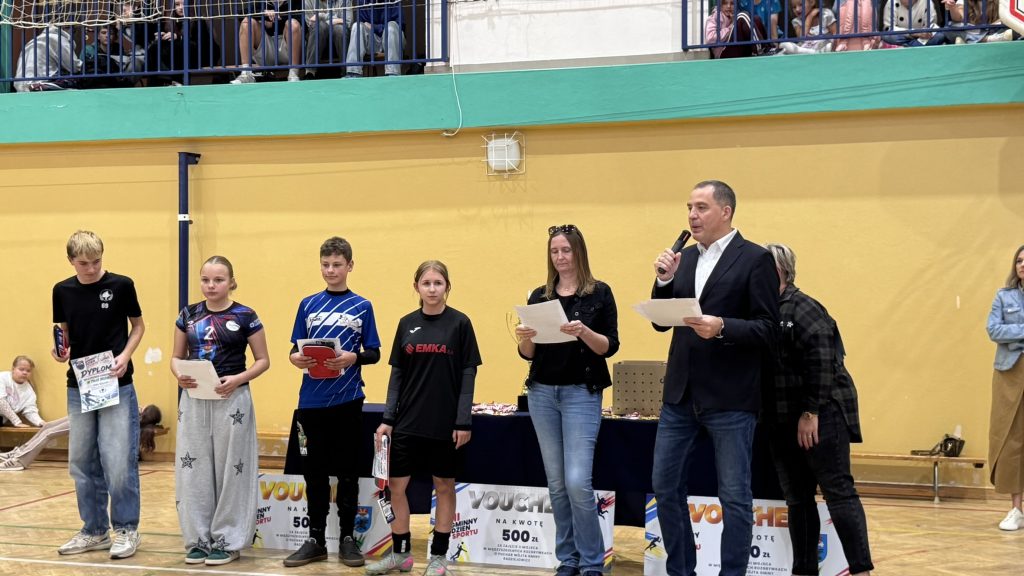 Grupa młodych sportowców i przedstawicieli organizatorów podczas wręczania nagród w hali sportowej. Uczestnicy stoją przed stołem z pucharami i dyplomami, a prowadzący trzyma mikrofon i odczytuje informacje z kartki.