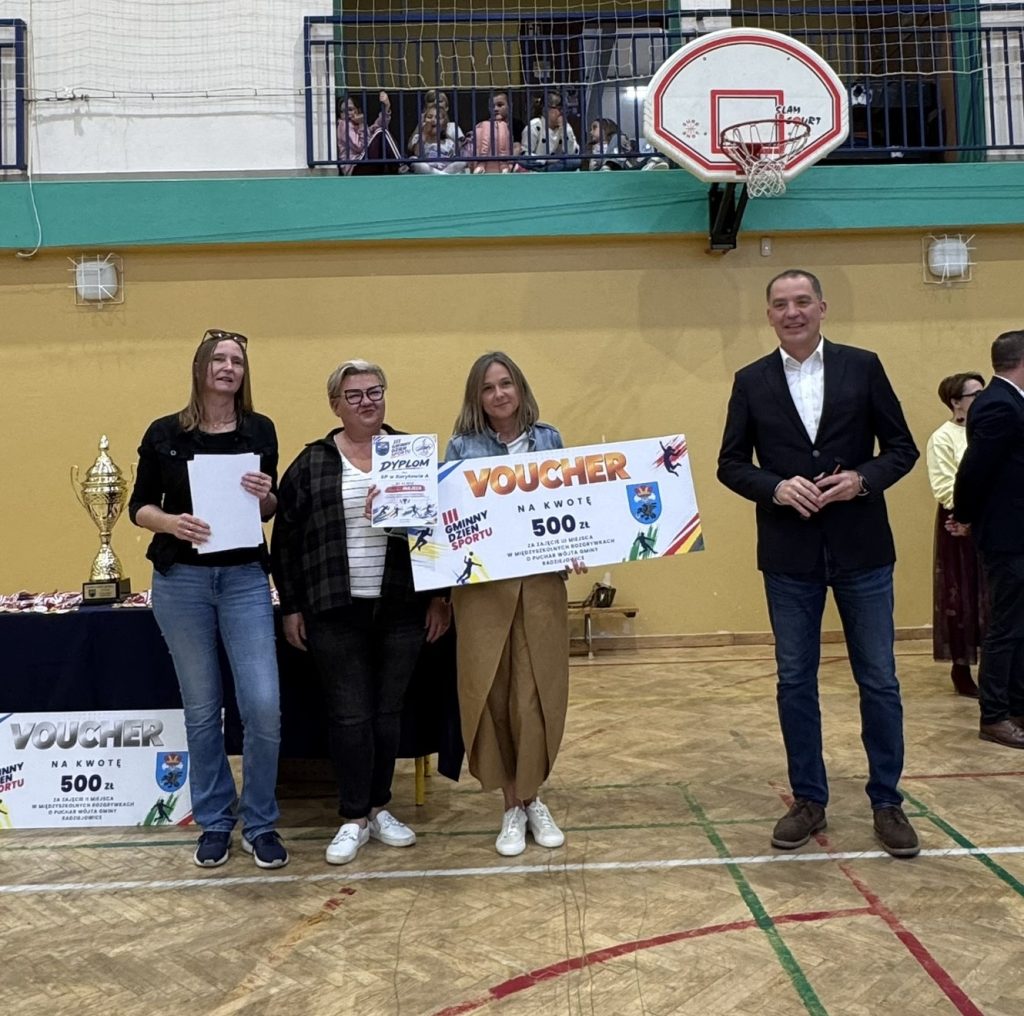 Przedstawiciele organizatorów pozują do zdjęcia z voucherem o wartości 500 zł, wręczonym w ramach Gminnego Dnia Sportu. W tle widoczna jest hala sportowa z koszem do koszykówki.