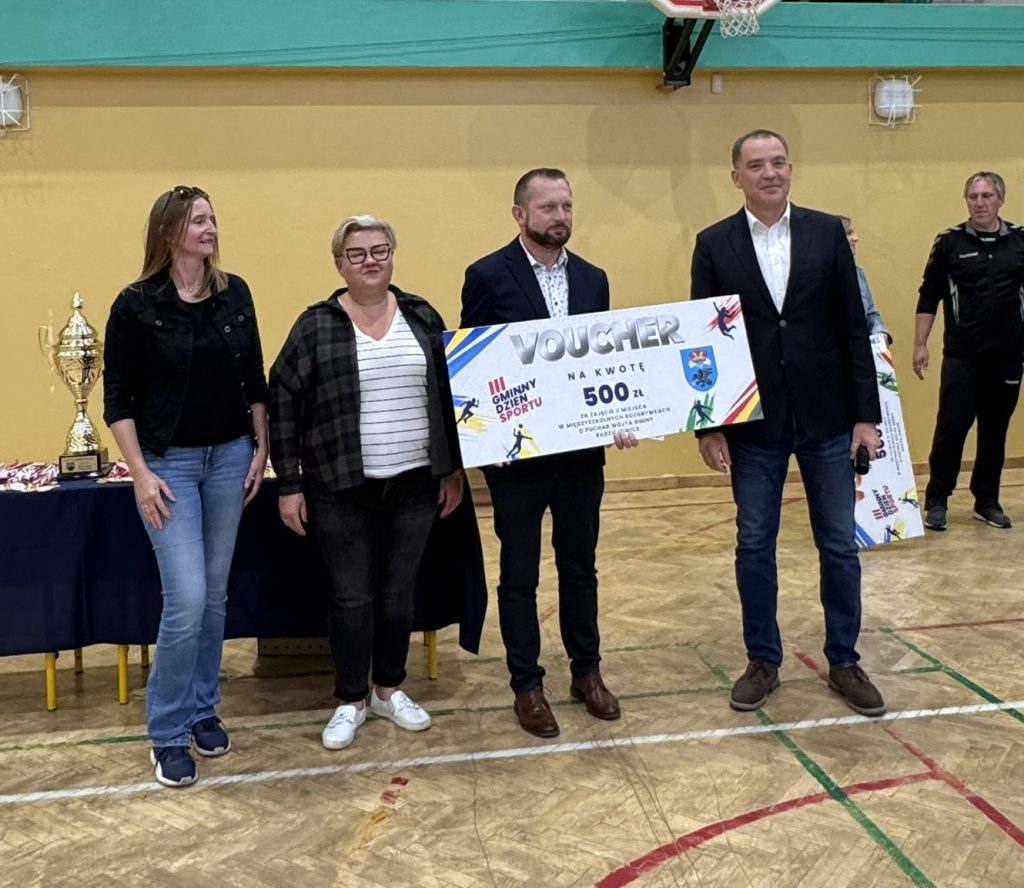 Przedstawiciele organizatorów oraz uczestnicy wydarzenia prezentują voucher o wartości 500 zł. Jedna z osób trzyma dużą planszę z napisem „Voucher”, a obok widoczny jest puchar.