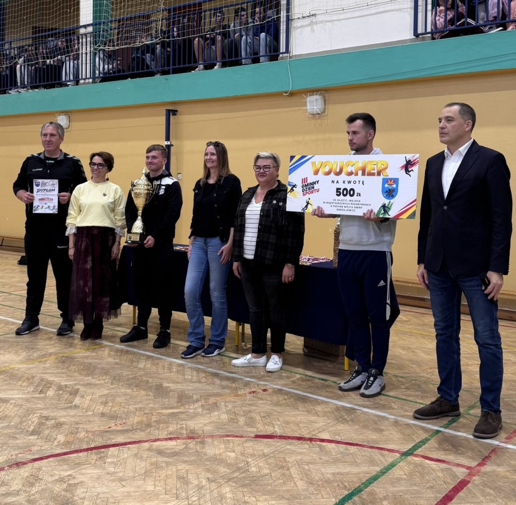 Grupa młodych sportowców w żółtych koszulkach oraz pozostali uczestnicy wydarzenia pozują do wspólnego zdjęcia. Dzieci prezentują zdobyte medale i dyplomy. Zdjęcie wykonano w hali sportowej.
