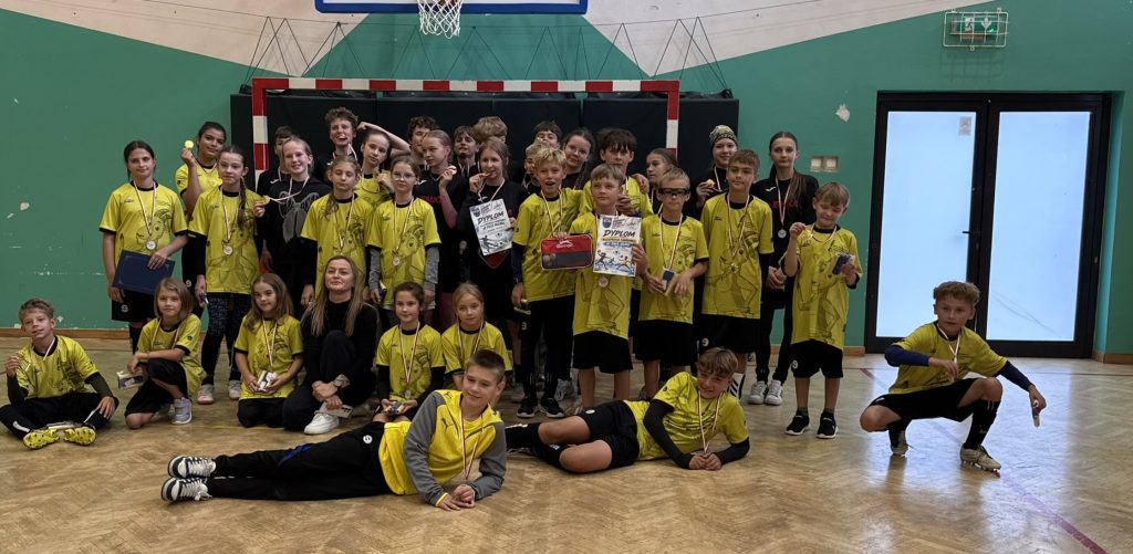 Grupa dzieci i młodzieży w strojach sportowych pozuje do wspólnego zdjęcia na tle bramki sportowej. Uczestnicy prezentują medale, dyplomy i puchar zdobyte podczas zawodów.