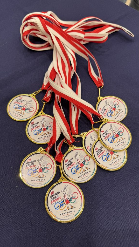 Zbliżenie na złote medale sportowe zawieszone na czerwono-białych wstążkach. Na medalach widoczne są oznaczenia Gminnego Dnia Sportu oraz nazwa miejscowości Korytów A.