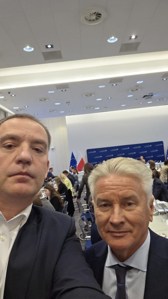 Dwóch mężczyzn podczas posiedzenia komisji wspólnej rządu i samorządu. W tle widoczni są uczestnicy spotkania oraz flagi Polski i Unii Europejskiej.