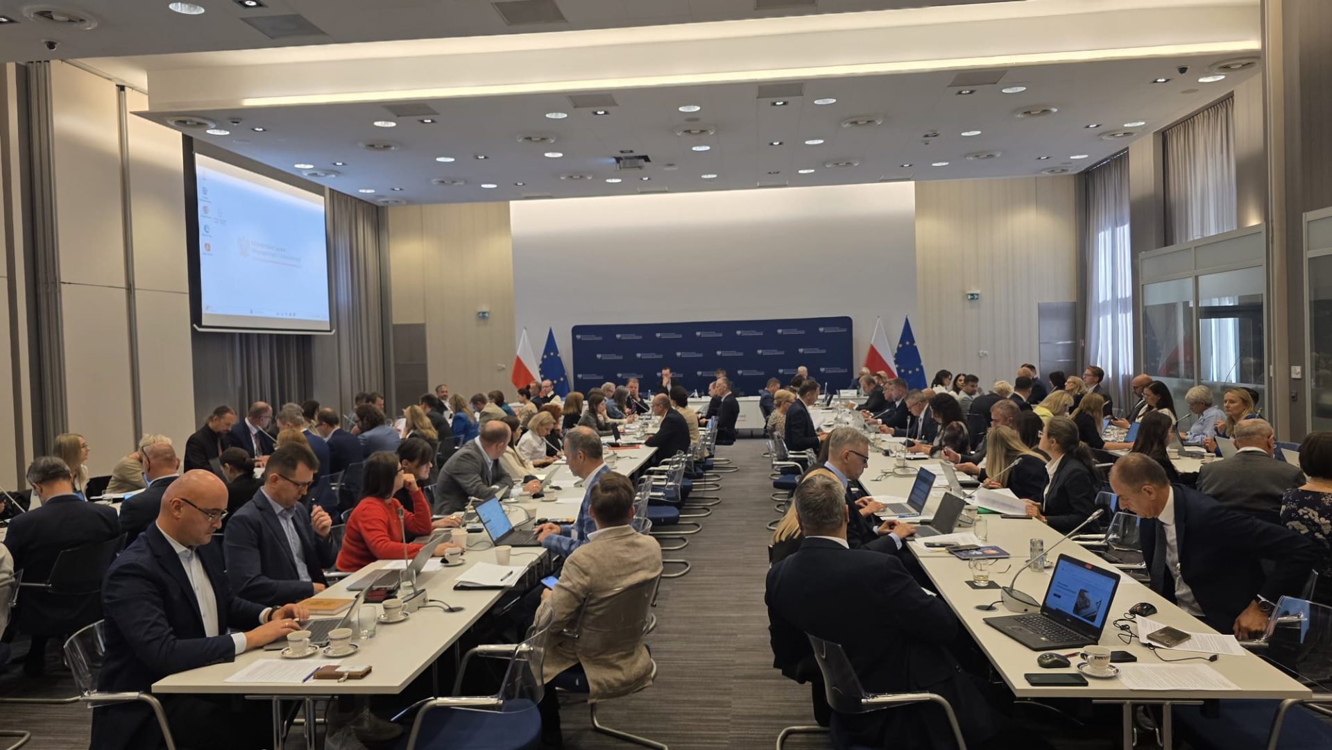 Posiedzenie komisji wspólnej rządu i samorządu w sali konferencyjnej Ministerstwa Spraw Wewnętrznych i Administracji. Przy stołach siedzą uczestnicy spotkania, a w tle widoczne są flagi Polski i Unii Europejskiej.