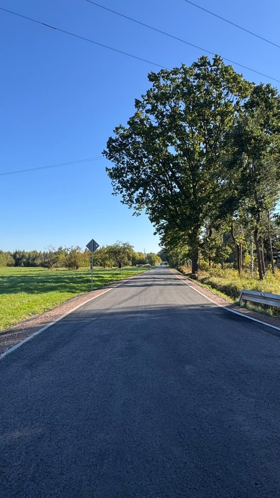 Nowo wyremontowana droga wśród terenów zielonych i drzew, z widocznym oznakowaniem drogowym oraz barierą ochronną.