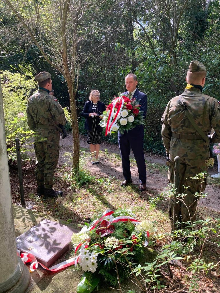 Mężczyzna w garniturze składający wieniec z czerwonych i białych kwiatów przy pomniku. Obok stoją żołnierze w mundurach, a w tle widoczna jest kobieta.