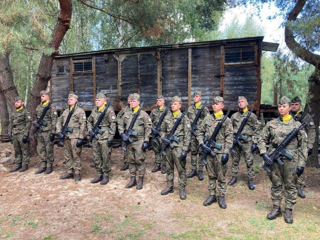 Szereg żołnierzy w mundurach wojskowych, z karabinami, stojących na baczność przed drewnianym budynkiem w otoczeniu drzew.