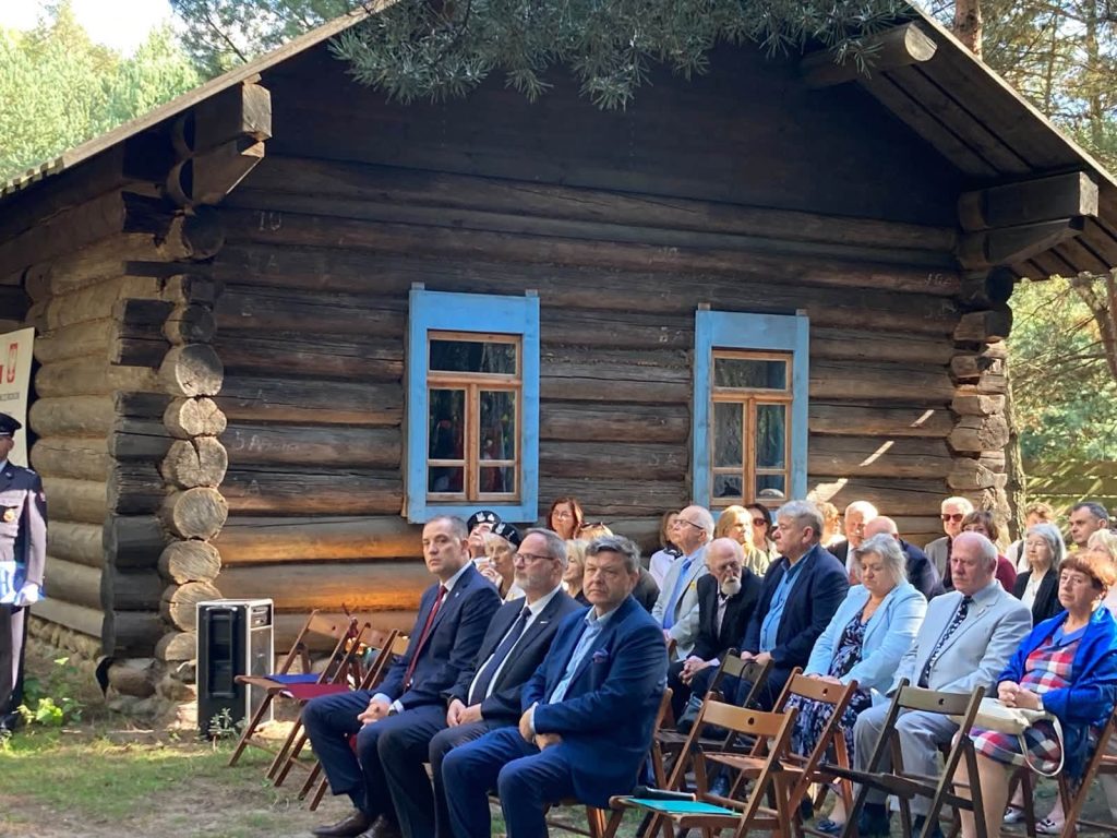 Uczestnicy uroczystości siedzący na krzesłach przed drewnianym budynkiem. W tle stoją poczty sztandarowe oraz żołnierze i przedstawiciele służb mundurowych.