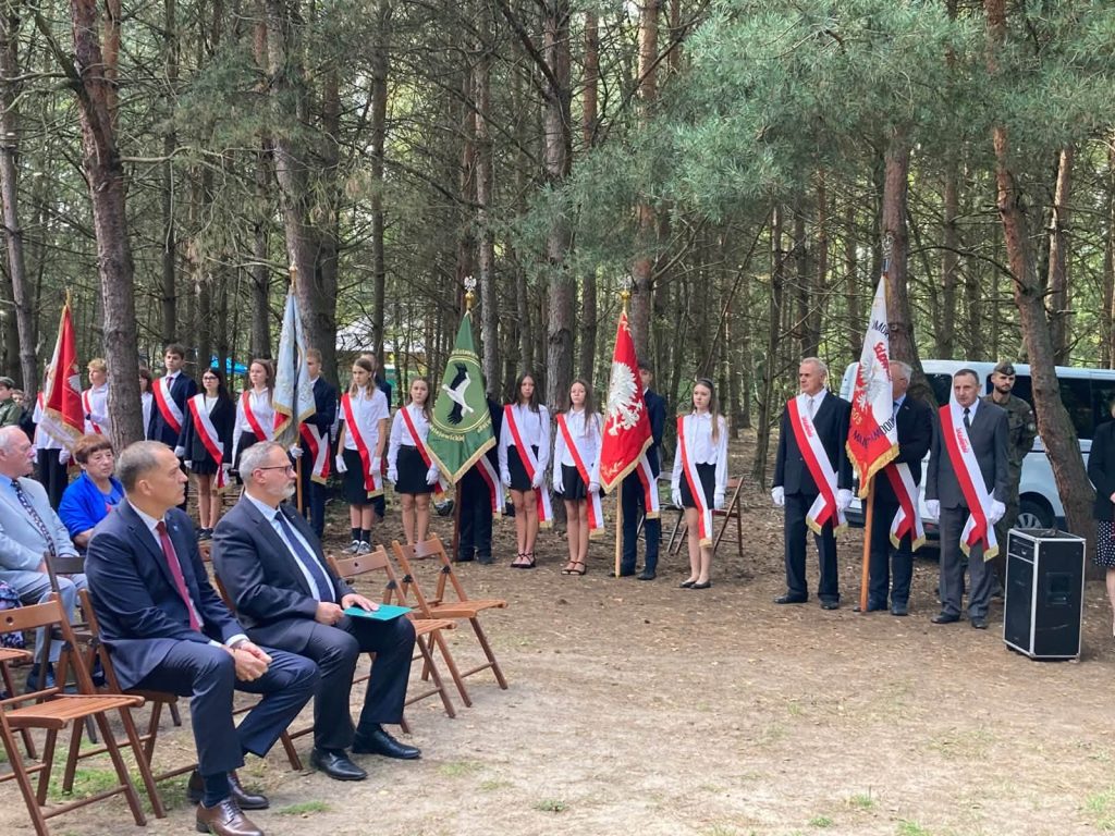 Uczestnicy uroczystości plenerowej siedzący na krzesłach oraz poczty sztandarowe i żołnierze stojący w szeregu w otoczeniu leśnym.