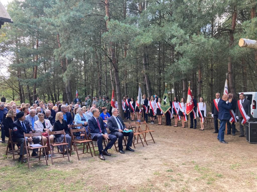 Uczestnicy uroczystości plenerowej siedzący na krzesłach oraz poczty sztandarowe i żołnierze stojący w szeregu w otoczeniu leśnym.