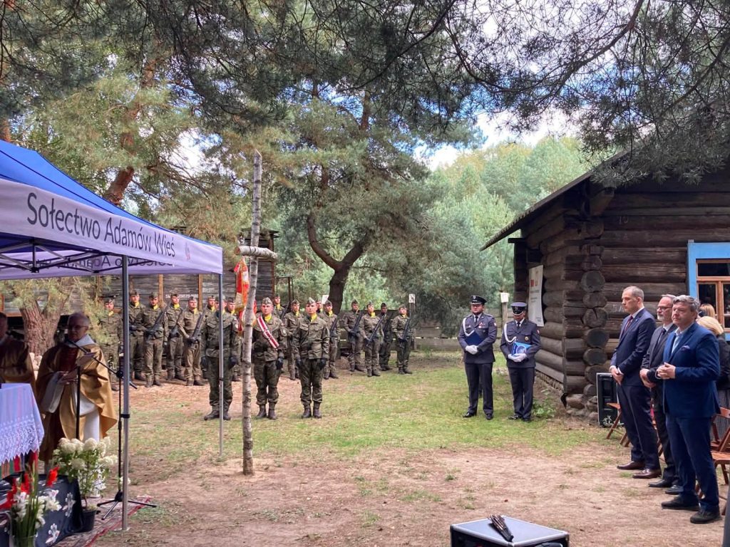 Uroczystość plenerowa przy drewnianym budynku. W centrum znajduje się mężczyzna przemawiający do mikrofonu, a za nim stoją żołnierze w mundurach wojskowych.