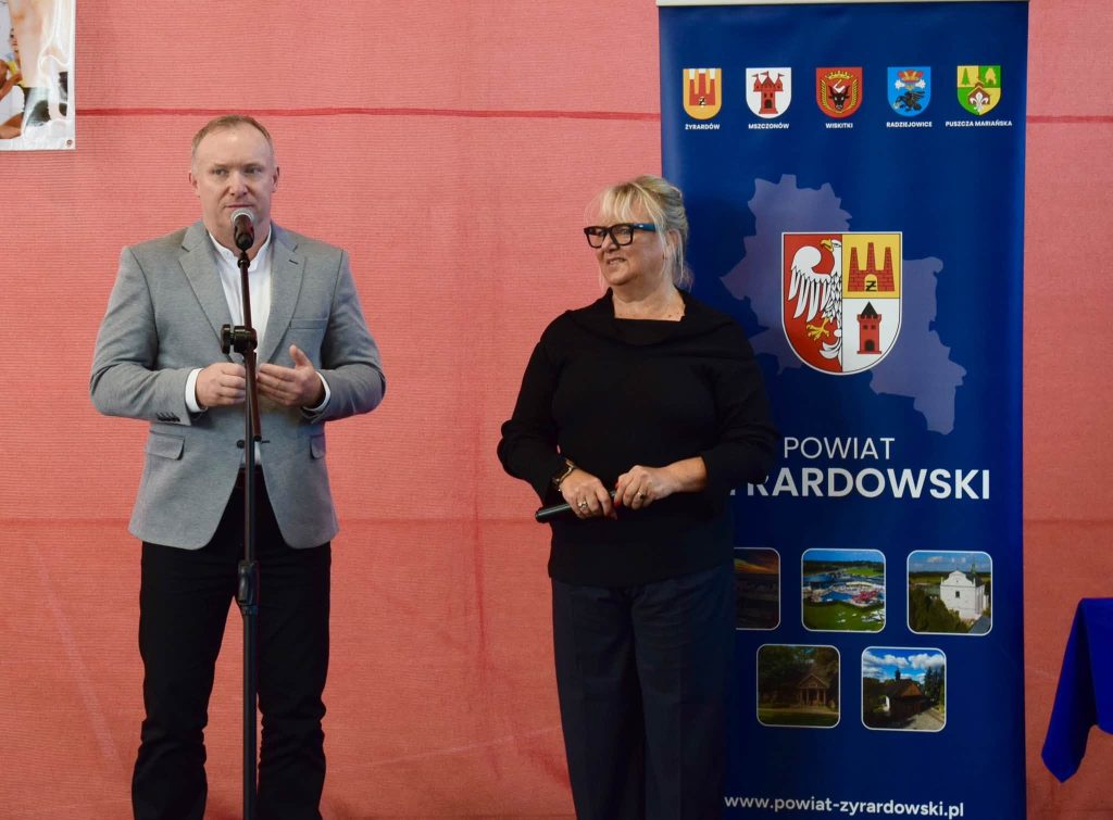Mężczyzna w szarej marynarce przemawia do mikrofonu podczas Gali Sportu Powiatu Żyrardowskiego. Obok niego stoi kobieta w czarnym stroju, a w tle widoczny jest niebieski baner z herbem i nazwą powiatu żyrardowskiego oraz herbami gmin.