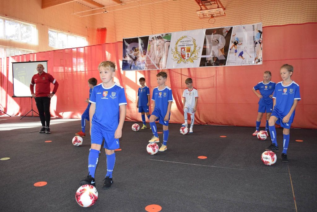 Chłopcy w niebieskich strojach sportowych oraz jeden zawodnik w białym stroju uczestniczą w pokazie piłkarskim podczas Gali Sportu Powiatu Żyrardowskiego. Każdy z nich ma piłkę przy nodze. Po lewej stronie stoi trener w czerwonej koszulce, a w tle widoczny jest baner wydarzenia i ekran projekcyjny.