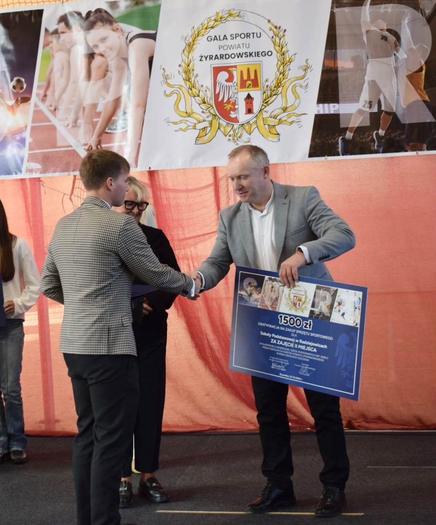 Mężczyzna w szarej marynarce wręcza młodemu mężczyźnie w kratkowanej marynarce tablicę z gratyfikacją w wysokości 1500 zł dla Szkoły Podstawowej w Radziejowicach za zajęcie I miejsca. W tle stoi kobieta w czarnym stroju, a nad nimi znajduje się baner Gali Sportu Powiatu Żyrardowskiego.