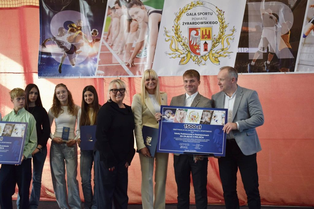 Grupa uczniów i przedstawicieli kadry szkolnej pozuje do wspólnego zdjęcia podczas Gali Sportu Powiatu Żyrardowskiego. Uczestnicy trzymają tablice z gratyfikacjami finansowymi i pamiątkowe dyplomy. Na środku stoją przedstawiciele organizatorów, a po prawej mężczyzna w szarej marynarce.