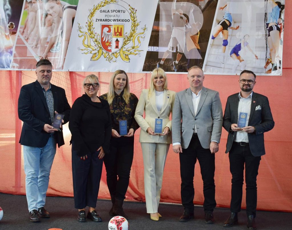 . Pięć osób pozuje do zdjęcia na tle baneru Gali Sportu Powiatu Żyrardowskiego. Trzymają pamiątkowe statuetki. Wśród nich znajdują się kobiety w czarnym i jasnym garniturze oraz mężczyźni w eleganckich strojach. U dołu zdjęcia widać piłkę nożną.