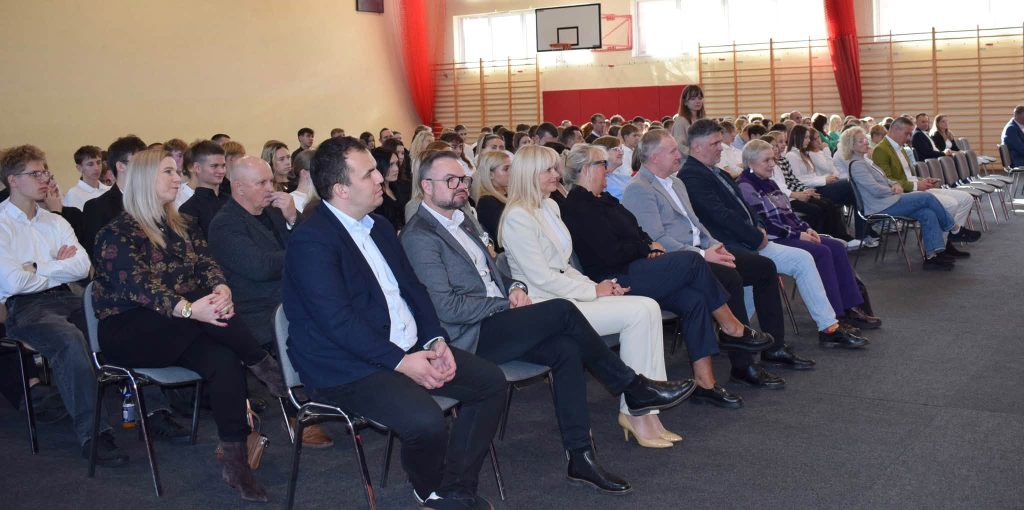 Uczestnicy Gali Sportu Powiatu Żyrardowskiego siedzą na krzesłach ustawionych w rzędach na sali gimnastycznej. W pierwszych rzędach znajdują się przedstawiciele władz i zaproszeni goście, a za nimi liczna grupa młodzieży. W tle widoczne są drabinki gimnastyczne, kosz do koszykówki i czerwone zasłony.