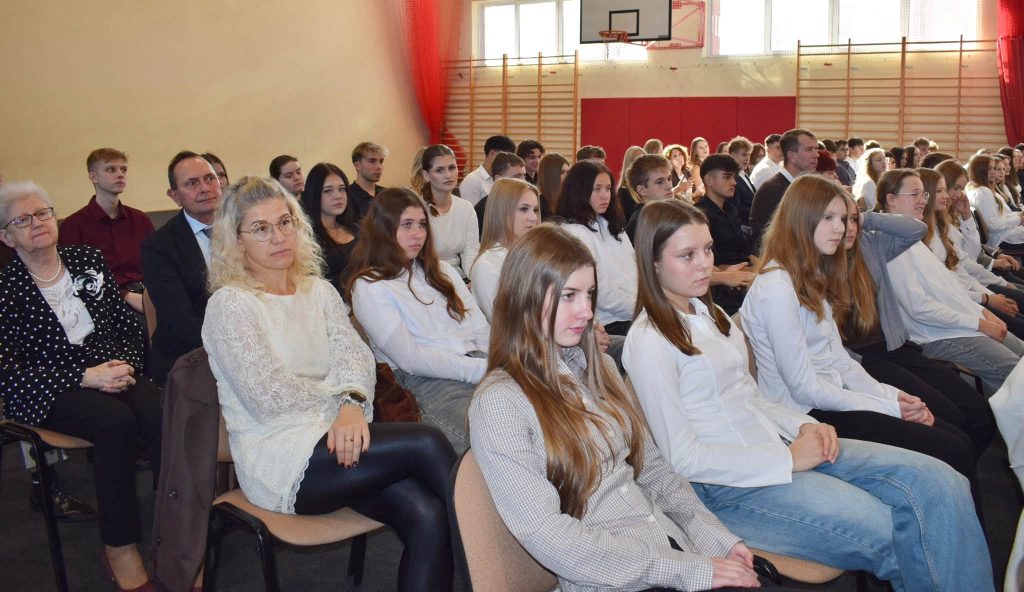 Uczestnicy Gali Sportu Powiatu Żyrardowskiego siedzą na krzesłach ustawionych w rzędach. Na pierwszym planie widać kobiety i dziewczęta, a w dalszych rzędach młodzież oraz dorosłych gości. W tle znajduje się sala gimnastyczna z drewnianymi drabinkami i czerwonymi zasłonami.