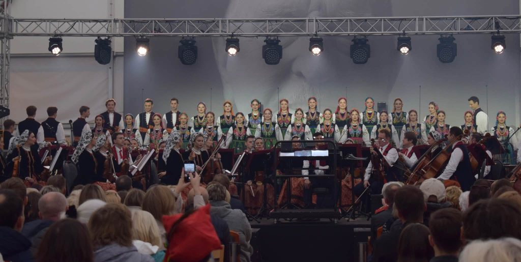 Zespół Pieśni i Tańca „Mazowsze” występuje na scenie wraz z orkiestrą. Artyści prezentują się w tradycyjnych strojach ludowych, a występ ogląda licznie zgromadzona publiczność.