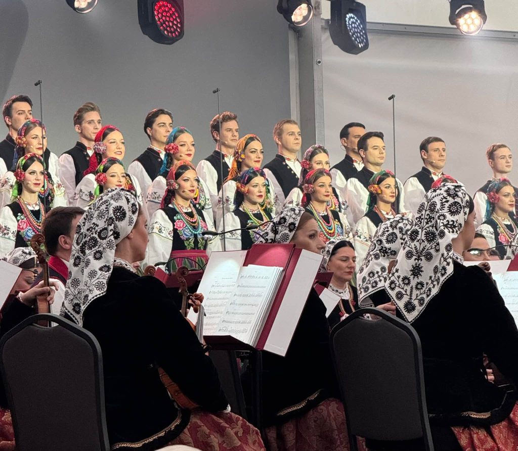 Artyści zespołu „Mazowsze” w tradycyjnych strojach ludowych podczas występu na scenie. W tle widoczny jest chór, a na pierwszym planie muzycy orkiestry z instrumentami smyczkowymi.