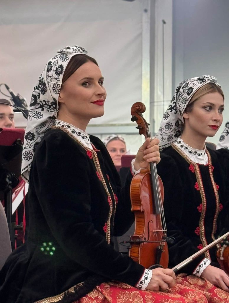 Dwie instrumentalistki zespołu „Mazowsze” w czarnych strojach ludowych, białych chustach z czarnym haftem i wzorzystych spódnicach. Jedna z kobiet trzyma skrzypce.