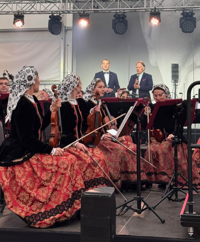 Muzycy orkiestry zespołu „Mazowsze” podczas koncertu. Na pierwszym planie widać instrumentalistki w tradycyjnych strojach ludowych, a w tle dwóch mężczyzn przemawiających na scenie.
