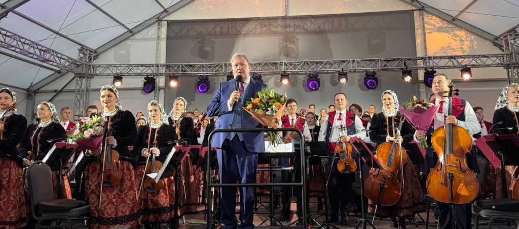 Mężczyzna w granatowym garniturze przemawia do publiczności ze sceny, trzymając mikrofon i bukiet kwiatów. Wokół niego stoją muzycy orkiestry zespołu „Mazowsze”.