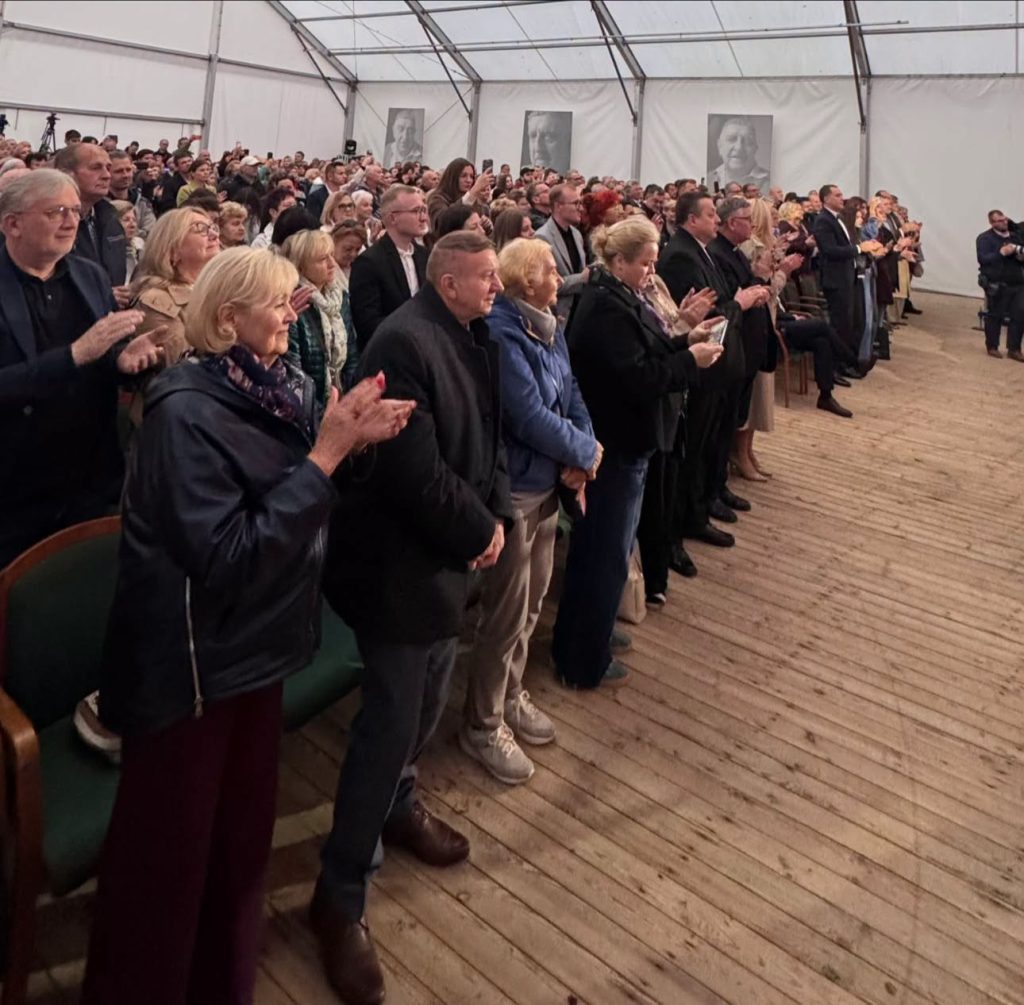 Publiczność zgromadzona na koncercie zespołu „Mazowsze” wstaje i nagradza artystów brawami. W tle widoczne są portrety oraz wnętrze namiotu koncertowego.