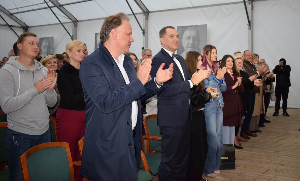Uczestnicy koncertu zespołu „Mazowsze” stoją i biją brawo artystom. Na pierwszym planie widać mężczyznę w granatowym płaszczu oraz gości ubranych w eleganckie stroje.