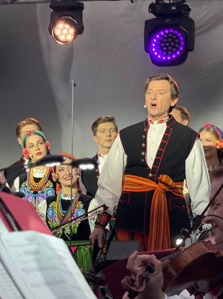 : Solista zespołu „Mazowsze” śpiewa na scenie w tradycyjnym stroju ludowym. Za nim stoją członkowie chóru w barwnych strojach regionalnych.