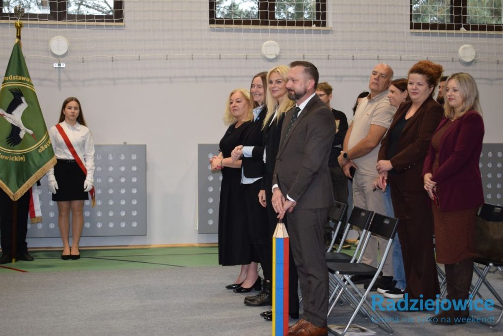 Przedstawiciele szkoły i zaproszeni goście uczestniczą w uroczystości pasowania na ucznia. Obok stoi poczet sztandarowy z zielonym sztandarem szkoły.