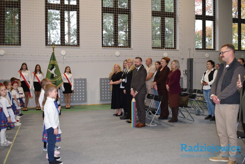 Uroczystość pasowania na ucznia w sali gimnastycznej. Na pierwszym planie stoi uczeń, a naprzeciwko niego dyrekcja i zaproszeni goście. W tle widoczni są pozostali uczniowie oraz poczet sztandarowy.