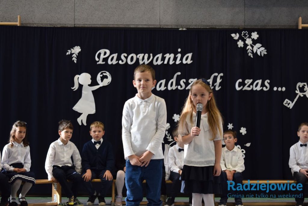 Dwoje dzieci występuje na szkolnej scenie przed dekoracją z napisem „Pasowania nadszedł czas”. Jedno z dzieci trzyma mikrofon, a pozostali uczniowie siedzą na ławkach w tle.