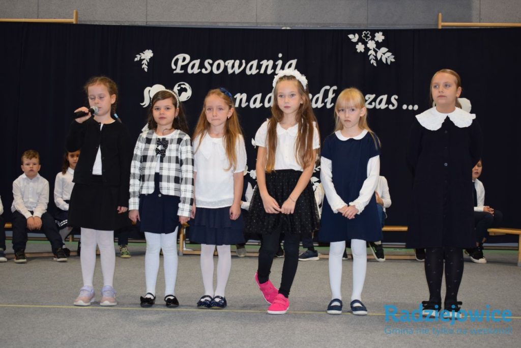 Grupa uczennic występuje przed publicznością podczas szkolnej uroczystości. Dziewczynki stoją w szeregu na tle dekoracji z napisem „Pasowania nadszedł czas”.