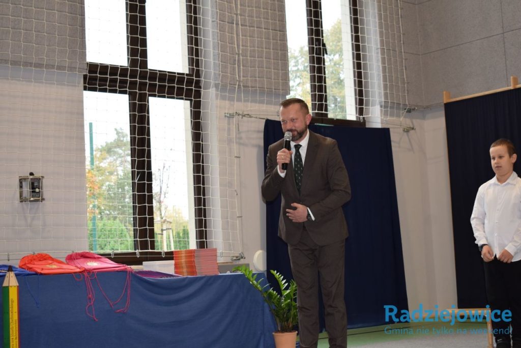 Dyrektor szkoły przemawia do uczestników uroczystości, trzymając mikrofon. Obok znajduje się stół z przygotowanymi upominkami dla uczniów.
