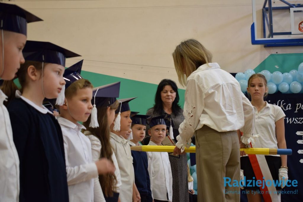 Nauczycielka trzyma żółty ołówek nad ramieniem stojącego przed nią ucznia podczas symbolicznego pasowania. W tle czekają pozostali pierwszoklasiści i nauczyciele.