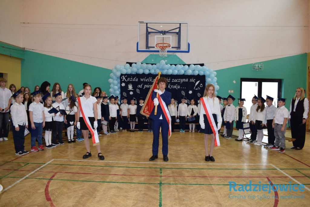 Poczet sztandarowy szkoły stoi na środku sali gimnastycznej. Uczniowie w biretach i odświętnych strojach ustawieni są po obu stronach, a w tle widoczna jest dekoracja z napisem „Każda wielka podróż zaczyna się od pierwszego kroku”.