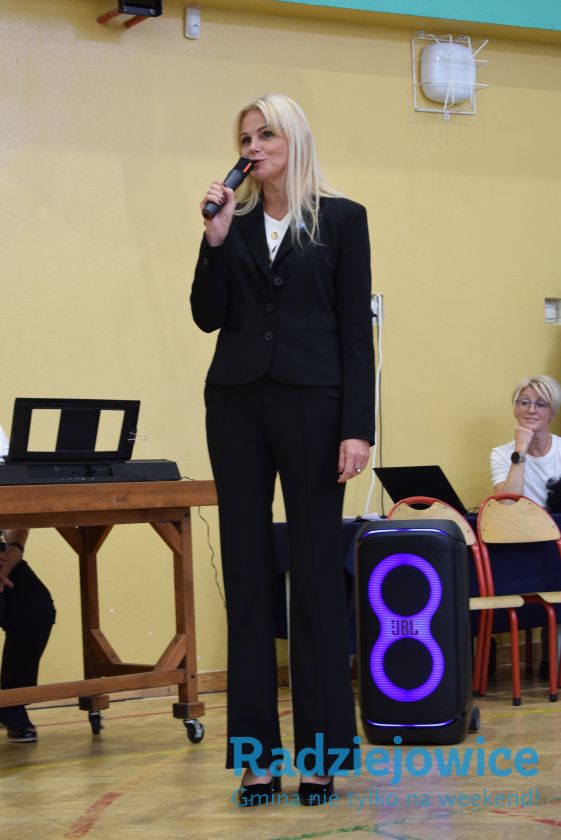 Kobieta w czarnym garniturze przemawia do uczestników uroczystości pasowania na pierwszoklasistów, trzymając mikrofon. Obok znajduje się głośnik i stół z laptopem.