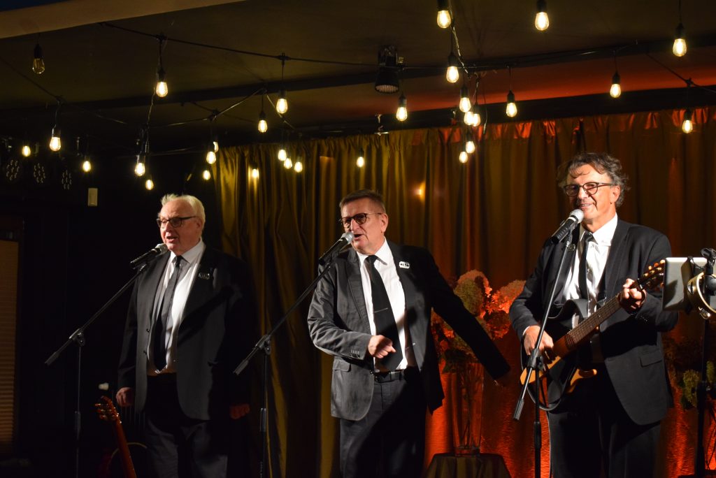 Trzech mężczyzn w eleganckich strojach występuje na scenie, śpiewając przy mikrofonach. Mężczyzna po prawej stronie gra na gitarze.
