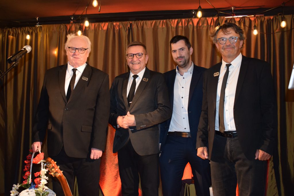 Czterech mężczyzn pozuje do zdjęcia na scenie. Trzech z nich ma na sobie ciemne garnitury i krawaty, a czwarty granatowy garnitur bez krawata.
