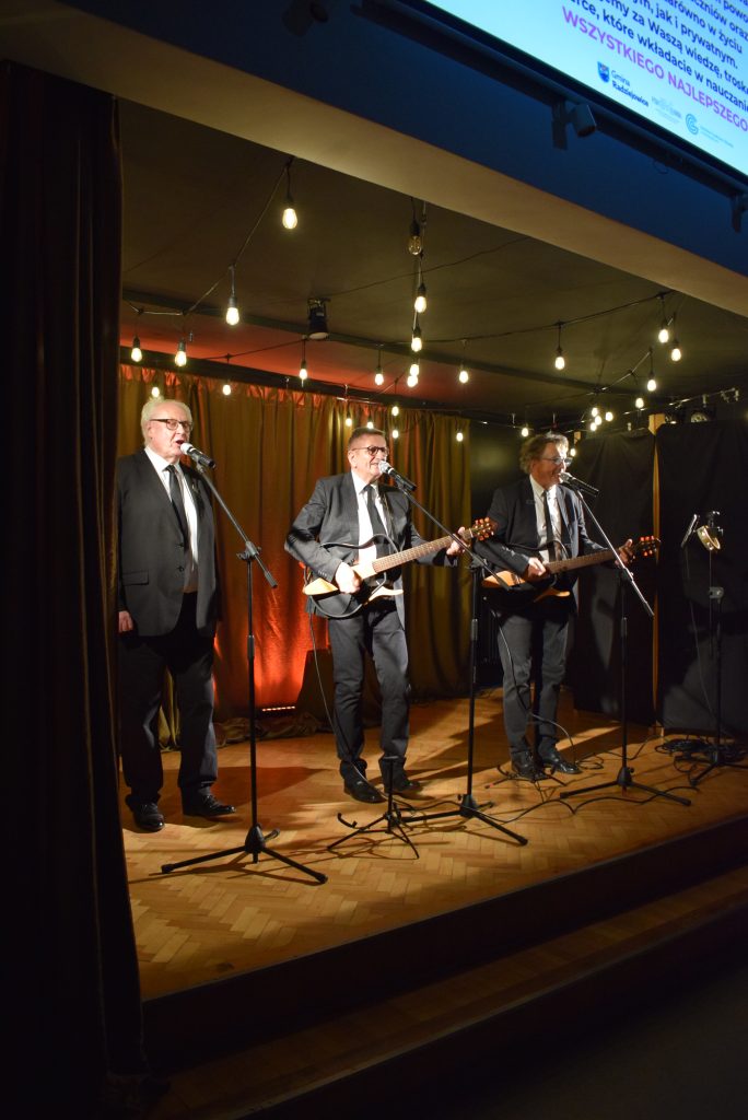 Trzech mężczyzn występuje na scenie przy mikrofonach. Dwóch gra na gitarach, a wszyscy są ubrani w eleganckie stroje. W tle widoczne są dekoracyjne oświetlenie i zasłony.