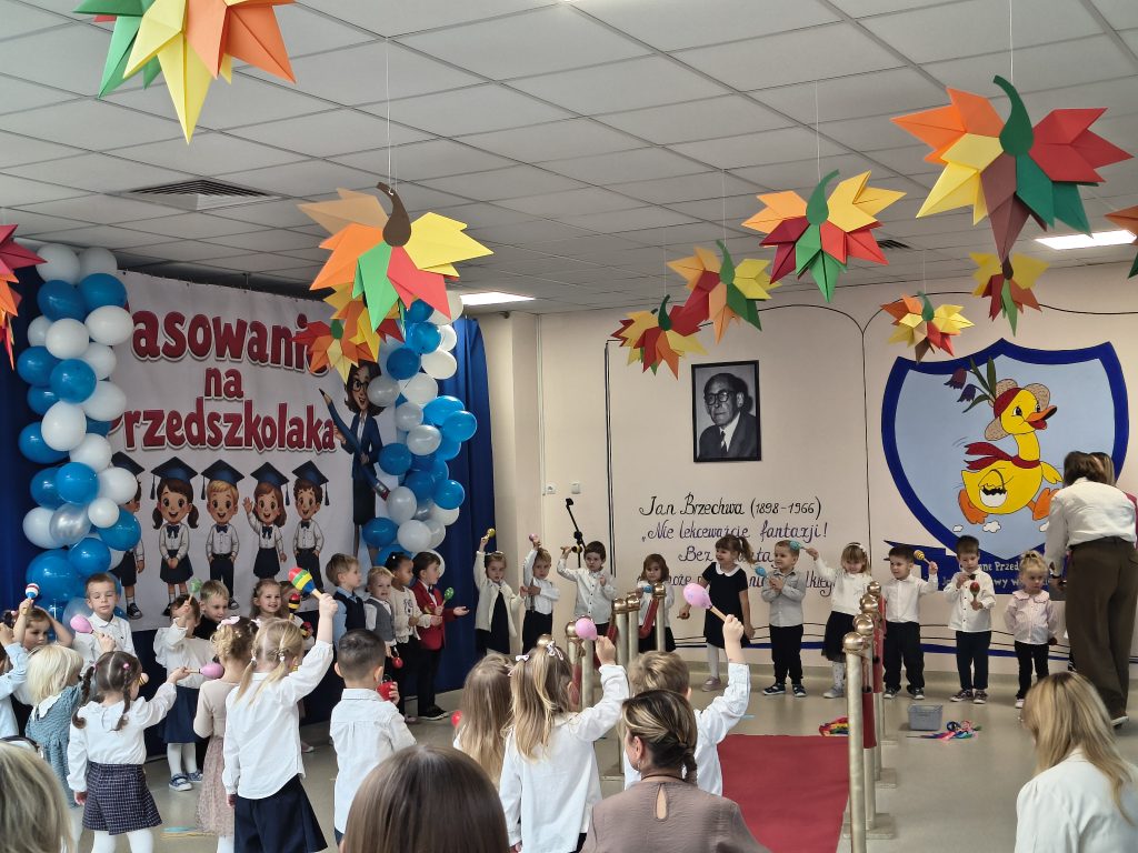 Grupa przedszkolaków stoi przed dekoracyjną ścianką podczas uroczystości pasowania. Dzieci unoszą kolorowe instrumenty i uczestniczą we wspólnym występie. W tle widoczne są niebiesko-białe balony, kolorowe dekoracje oraz czerwony dywan.